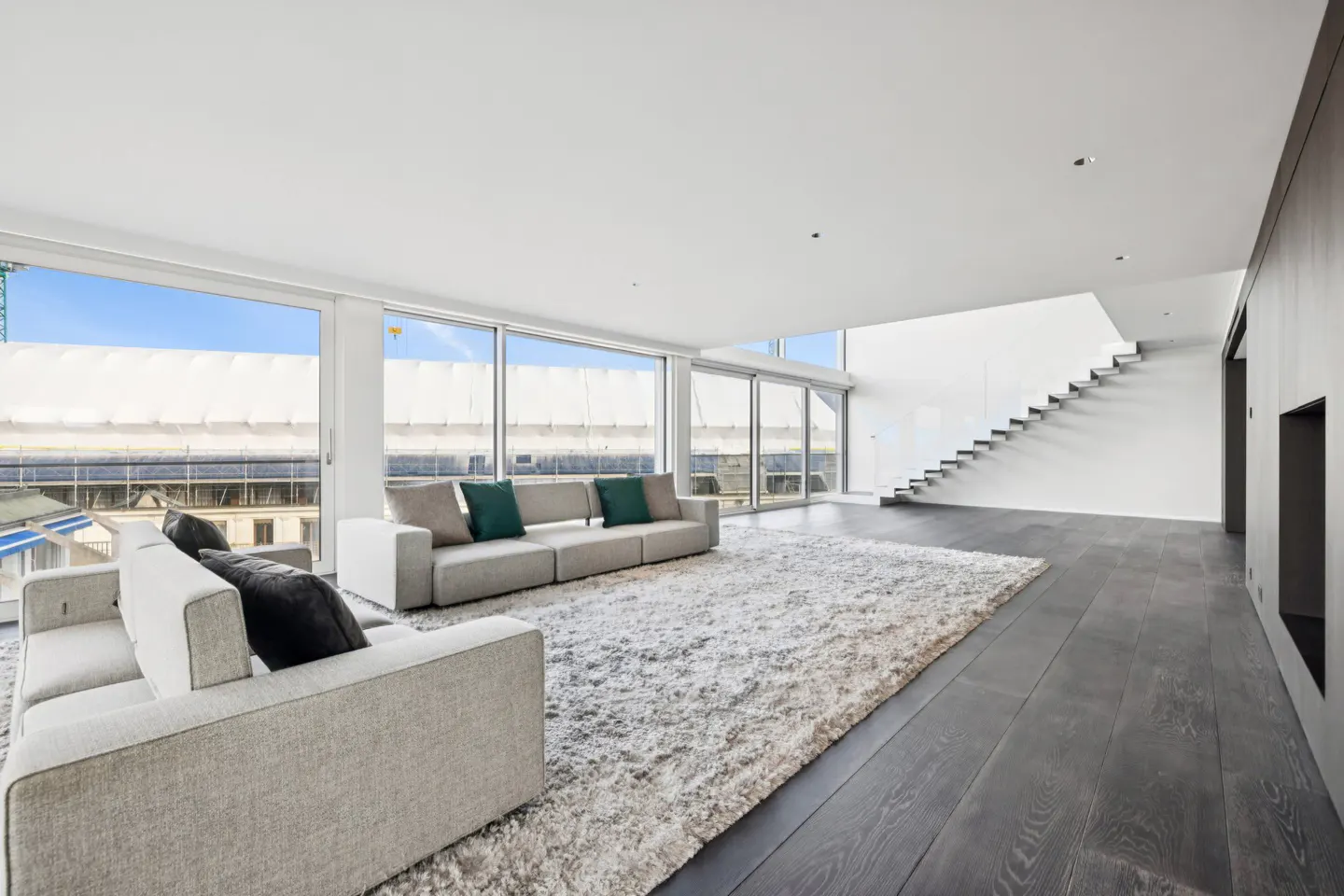 Bright, modern living room with gray sofas, green pillows, and a white shag rug on dark wood floors. Large windows offer a city view. A staircase is visible.