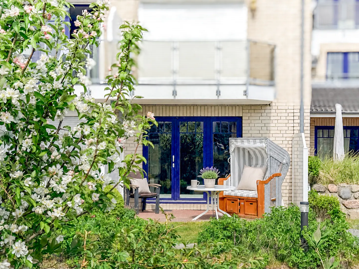 Patio with blue doors, brick wall, and blooming tree. Wicker chair, table, and chair on the patio. Greenery in the foreground.