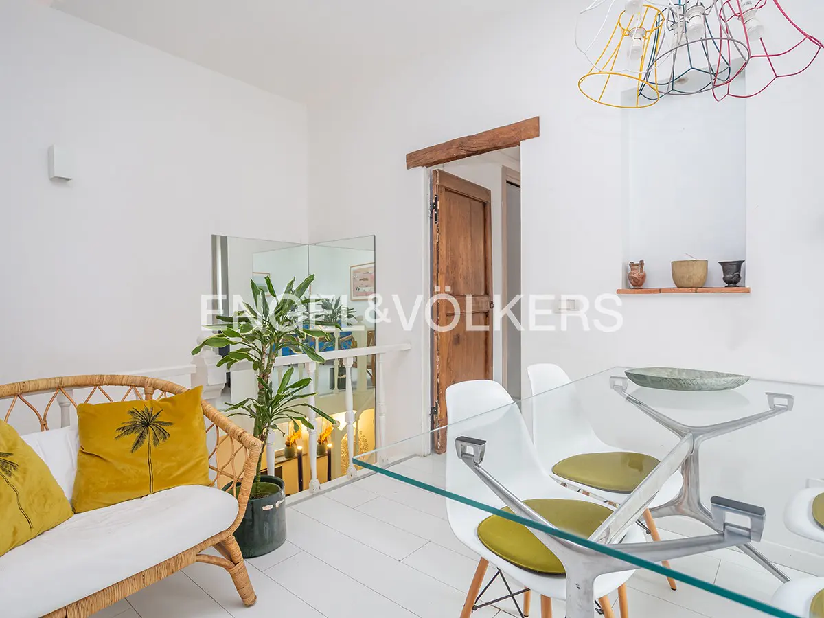 Bright dining area with white walls, a glass table with white chairs, and a rattan sofa with yellow pillows. A plant sits near a mirrored wall.
