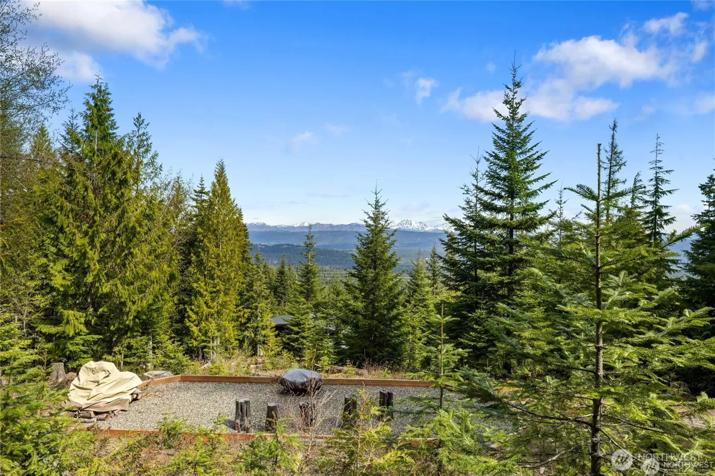 Scenic view of a gravel patio surrounded by tall green trees, with snow-capped mountains visible in the distance under a blue sky.