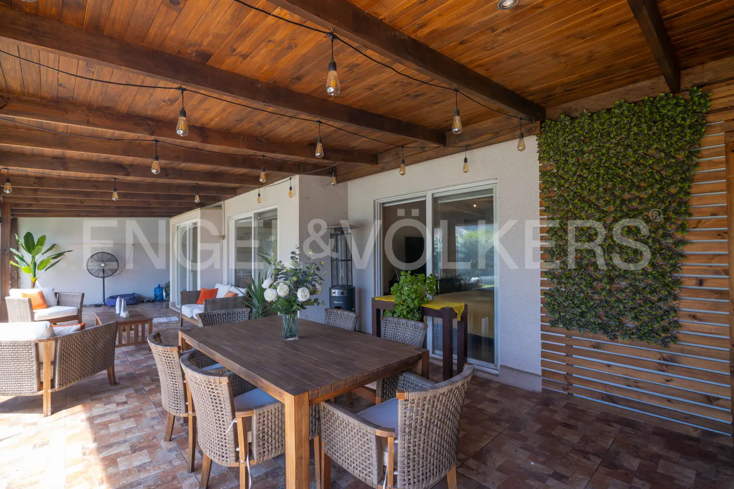 Outdoor patio with a wooden table, wicker chairs, and string lights under a wooden ceiling. Green wall decor adds a natural touch.