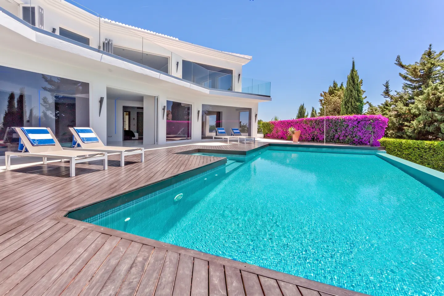 Luxury villa with a turquoise pool, wooden deck, and white lounge chairs. Bougainvillea and trees in the background under a clear blue sky.