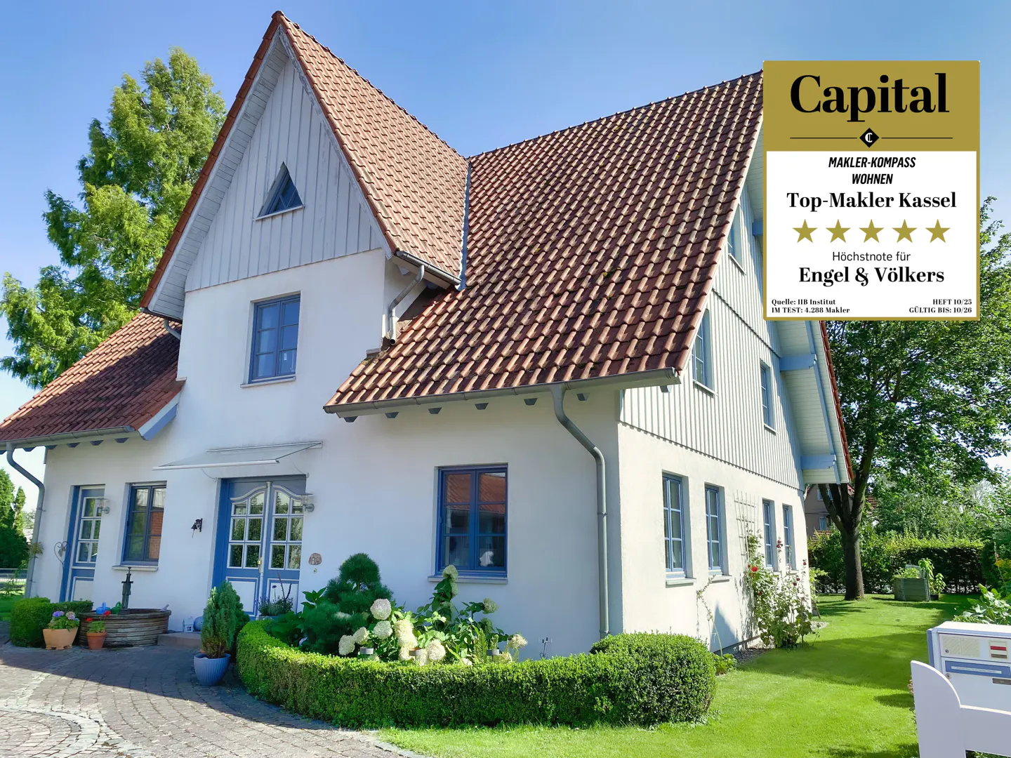 Two-story white house with a red tile roof, blue trim, and a manicured green lawn. "Capital" award logo is visible.
