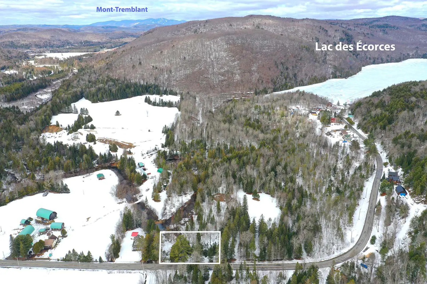 Aerial view of a snow-covered landscape with trees, a road, and a lake in the background. Mont-Tremblant is visible in the distance.