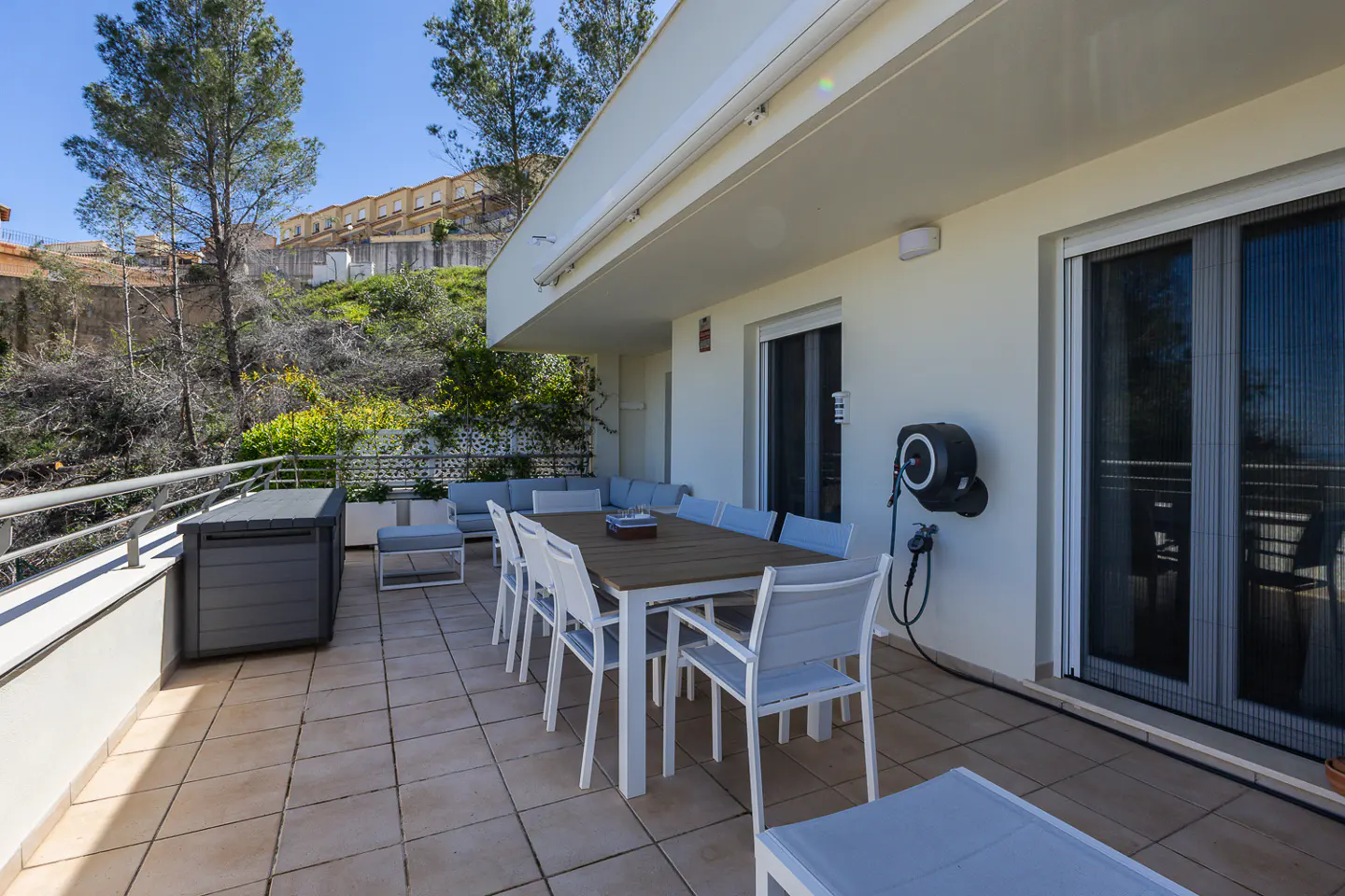 Outdoor patio with a large table and chairs, a gray storage box, and a view of a hillside with trees and buildings.