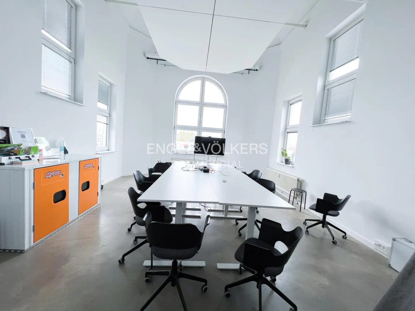 Bright, modern office space with white walls, large windows, and a long white conference table surrounded by black chairs. Orange storage units are visible.