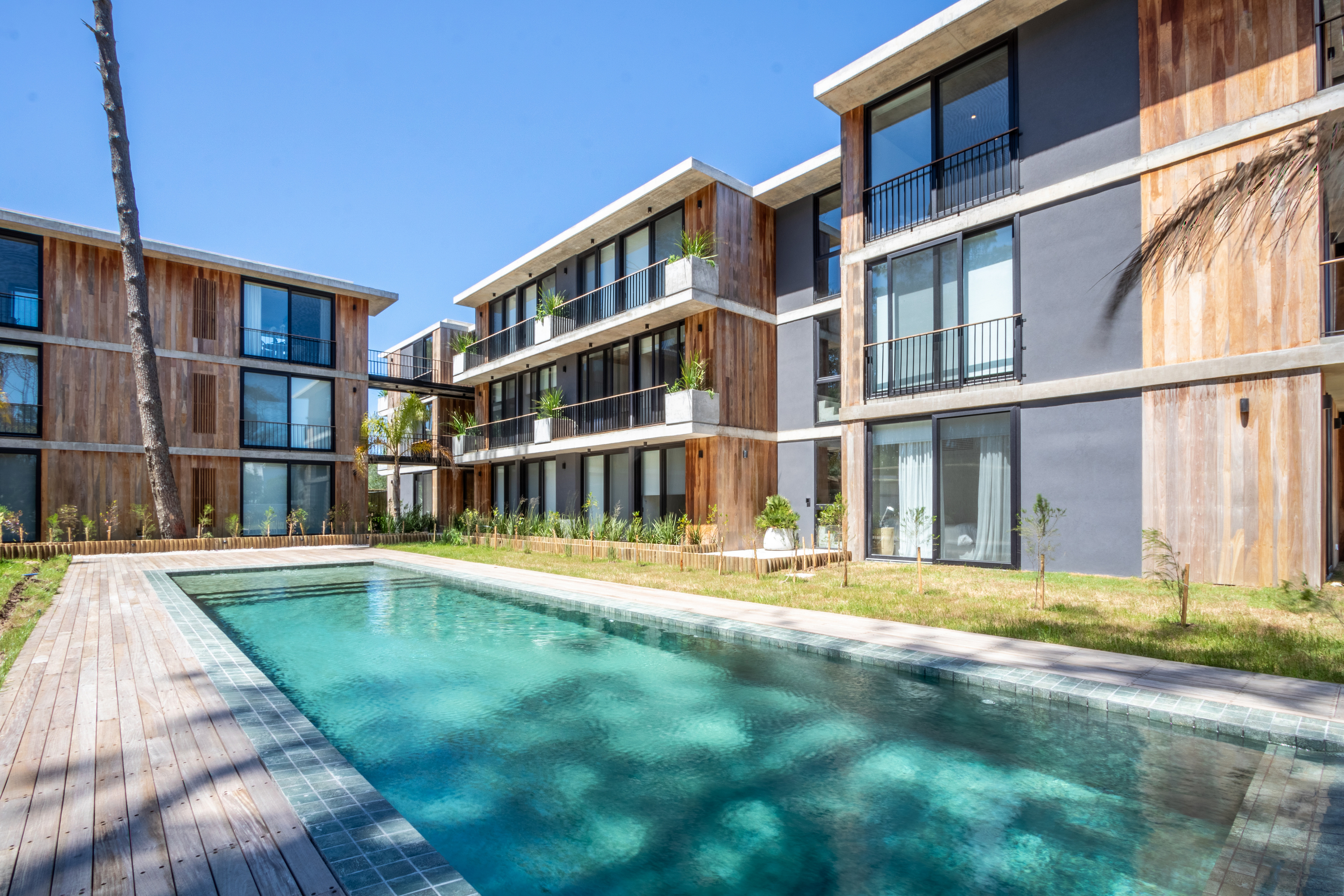 Modern apartments with a pool. The buildings have wood and gray exteriors, balconies, and large windows. The pool has a wooden deck.