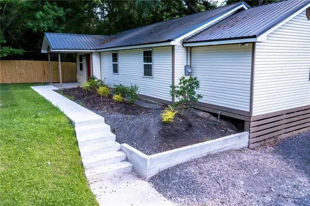 Exterior view of a one-story white house with a metal roof, a concrete walkway, and a small garden.