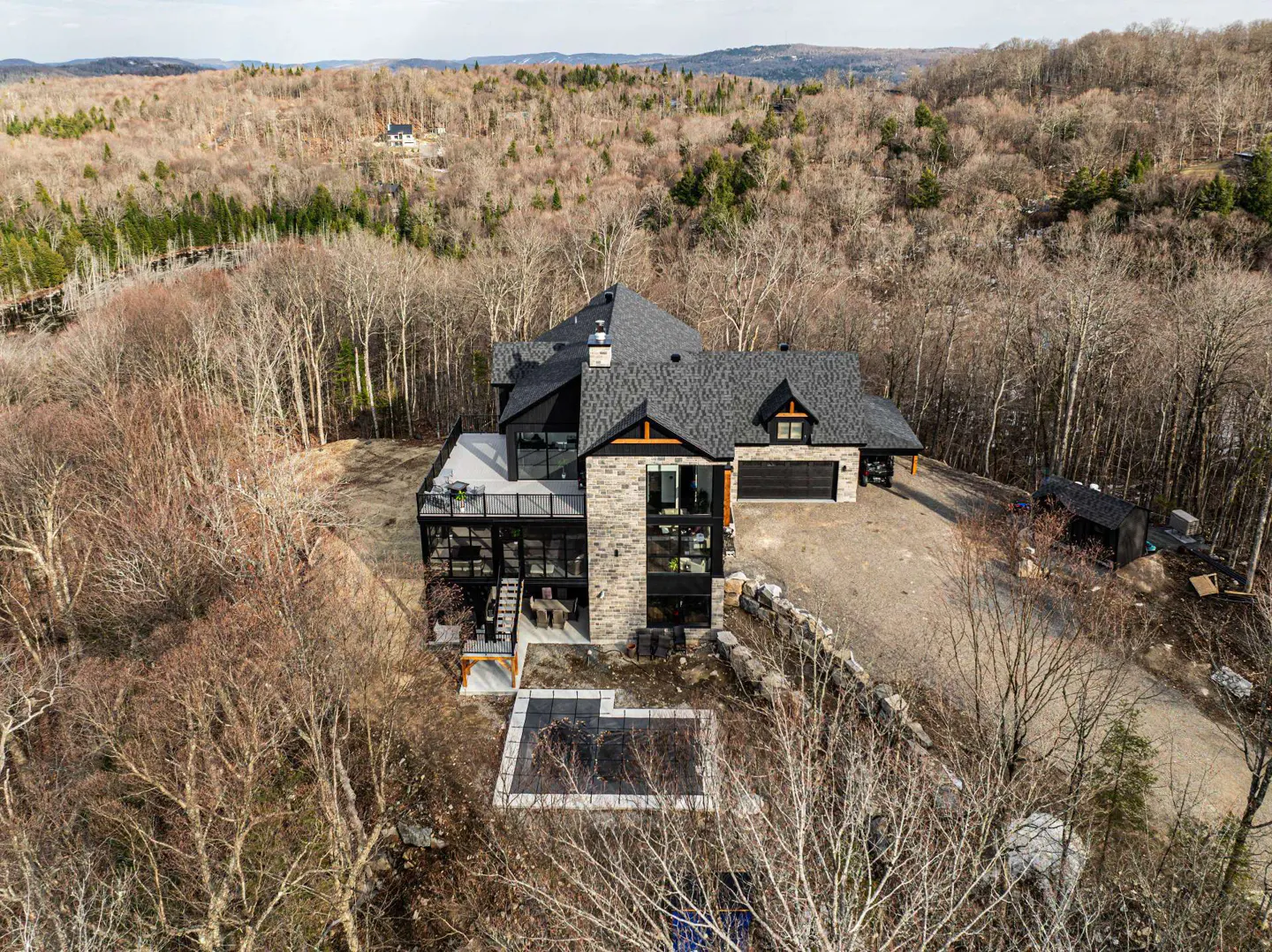 Aerial view of a modern stone house with a black roof, deck, pool, and forest background.