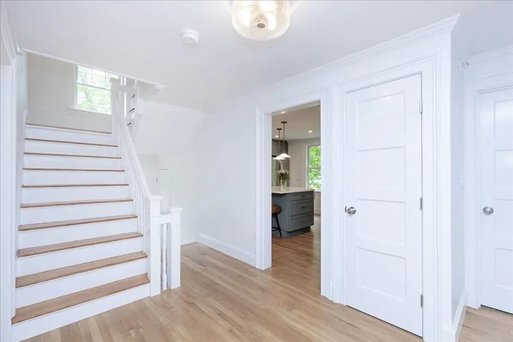 Bright foyer with hardwood floors, white walls, and staircase. A glimpse of the kitchen with a gray island is visible through an open doorway.
