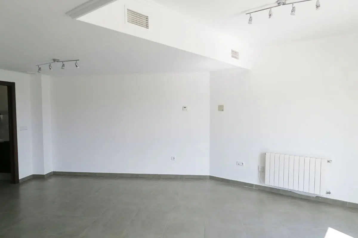 An empty room with white walls, gray tile floor, and track lighting. A white radiator is on the right wall. A doorway is visible on the left.