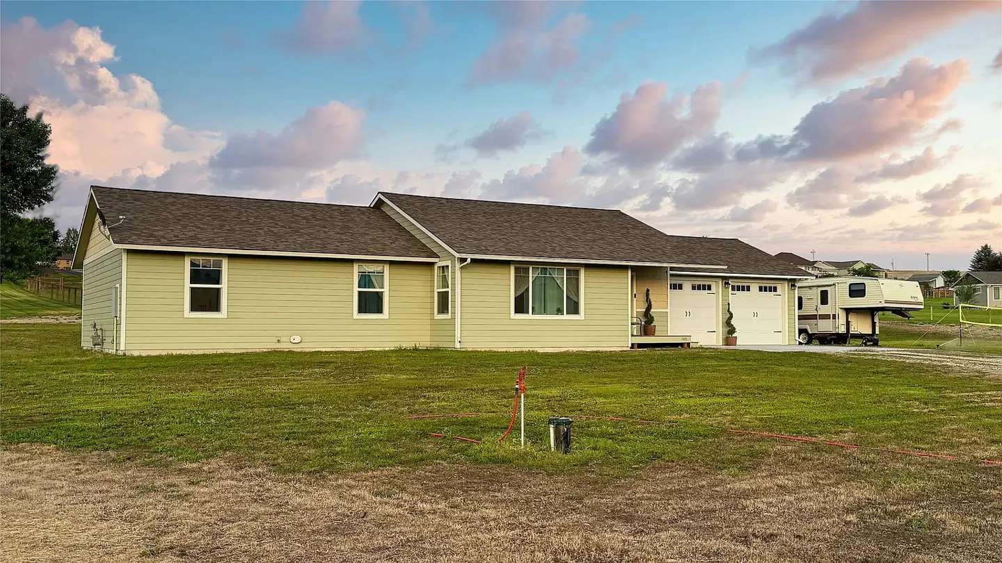 A light green single-story house with a brown roof, white garage doors, and a camper parked nearby.