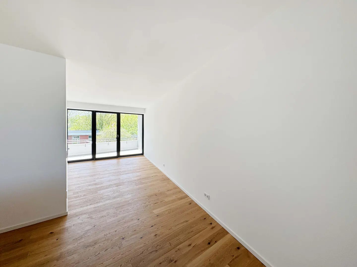 Empty room with white walls, light wood flooring, and large floor-to-ceiling windows overlooking a balcony and trees.