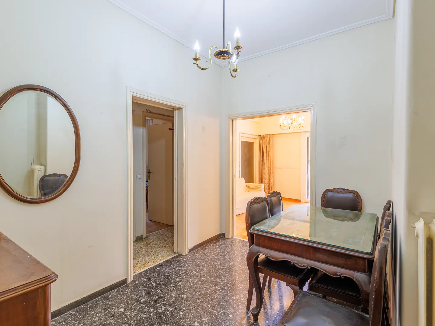 A room with a dining table, a mirror, and two doorways. The table is dark wood with a glass top and leather chairs. A chandelier hangs from the ceiling.