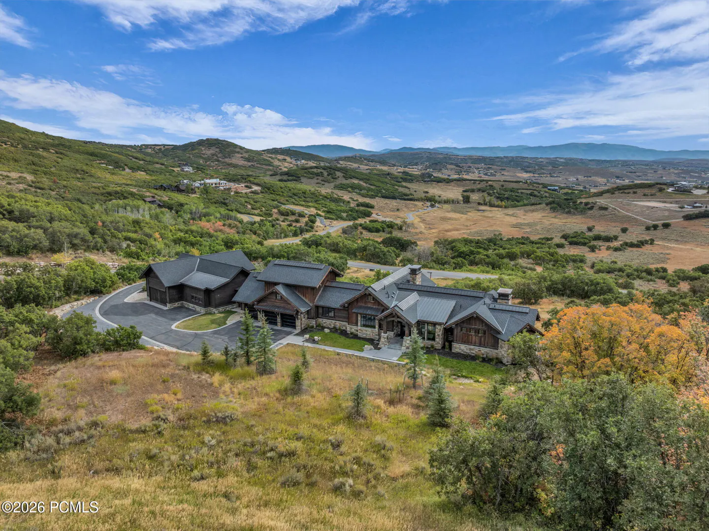 Aerial view of a large, rustic-style home with a dark roof, surrounded by green hills and a blue sky with scattered clouds.