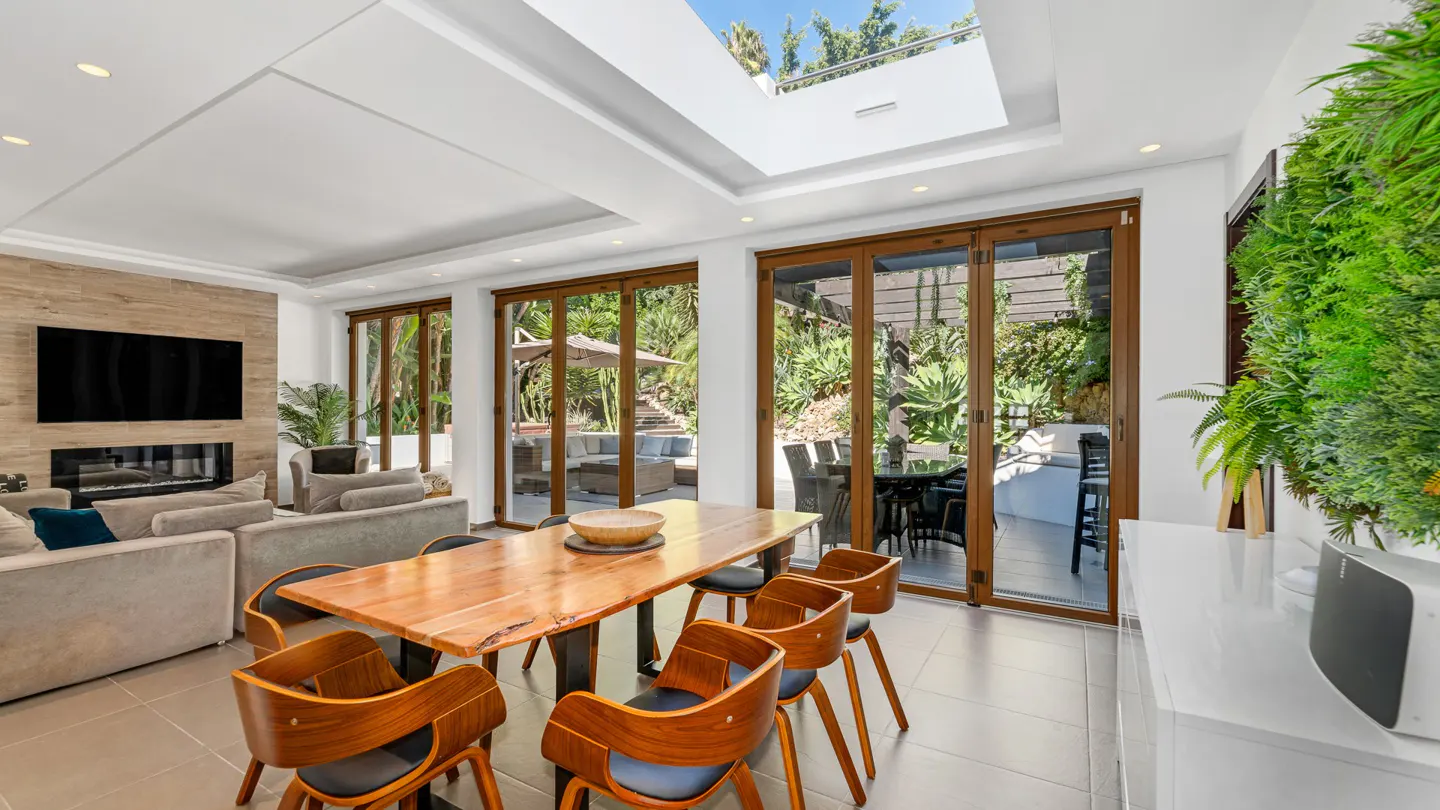 Bright, modern dining room with wood table, chairs, and folding doors to a lush outdoor patio. Skylight above.