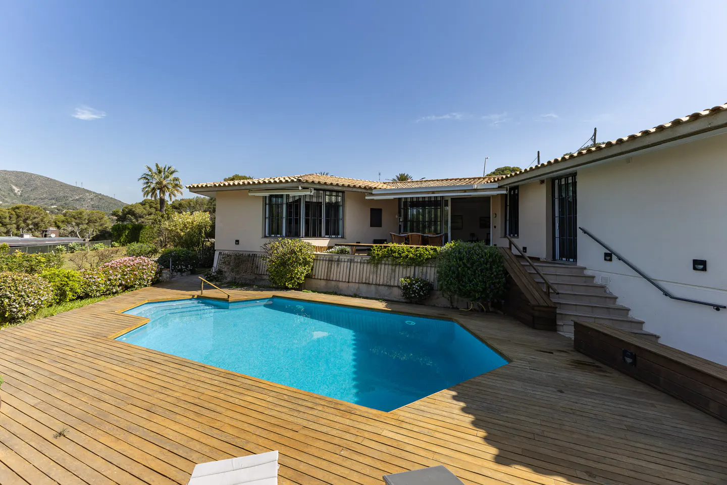 A backyard with a pool and a beige house with a brown roof, surrounded by a wooden deck and green plants.