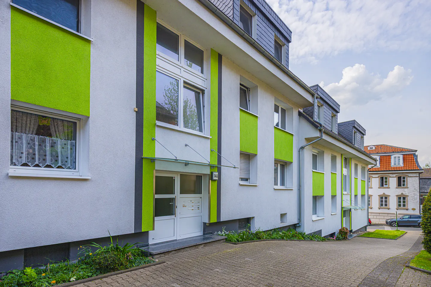 Exterior view of a modern apartment building with white walls and green accents, a paved driveway, and a cloudy sky.