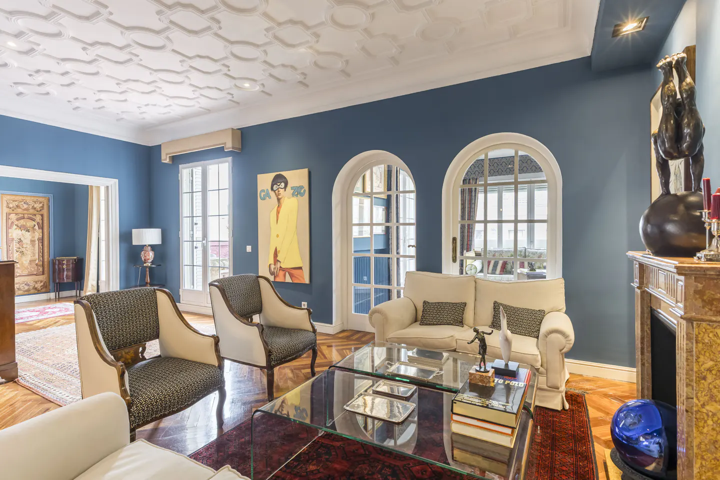 Living room with blue walls, white trim, and hardwood floors. Two chairs, a sofa, and a glass coffee table are arranged on a red rug. A painting hangs on the wall.