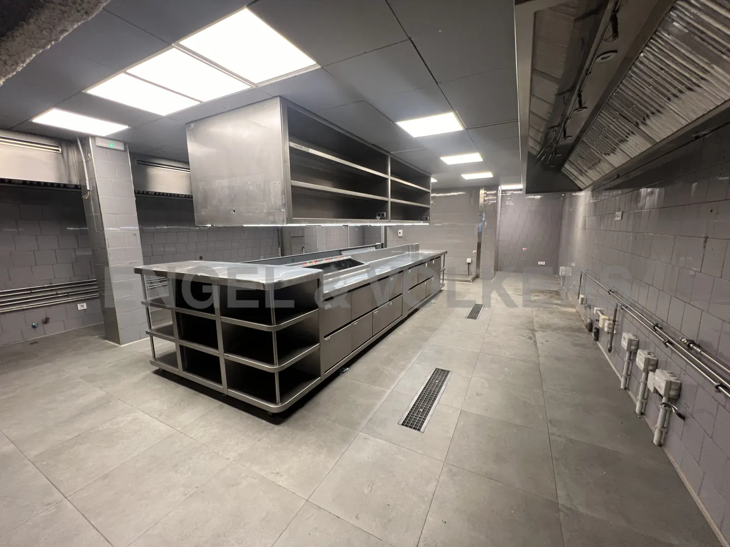 A commercial kitchen with stainless steel counters, shelves, and gray tiled walls and floors.