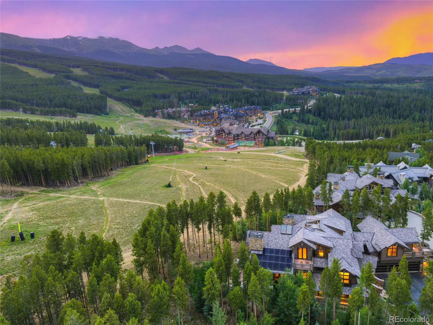 Aerial view of a large log cabin home surrounded by green trees, with mountains and a colorful sunset in the background.