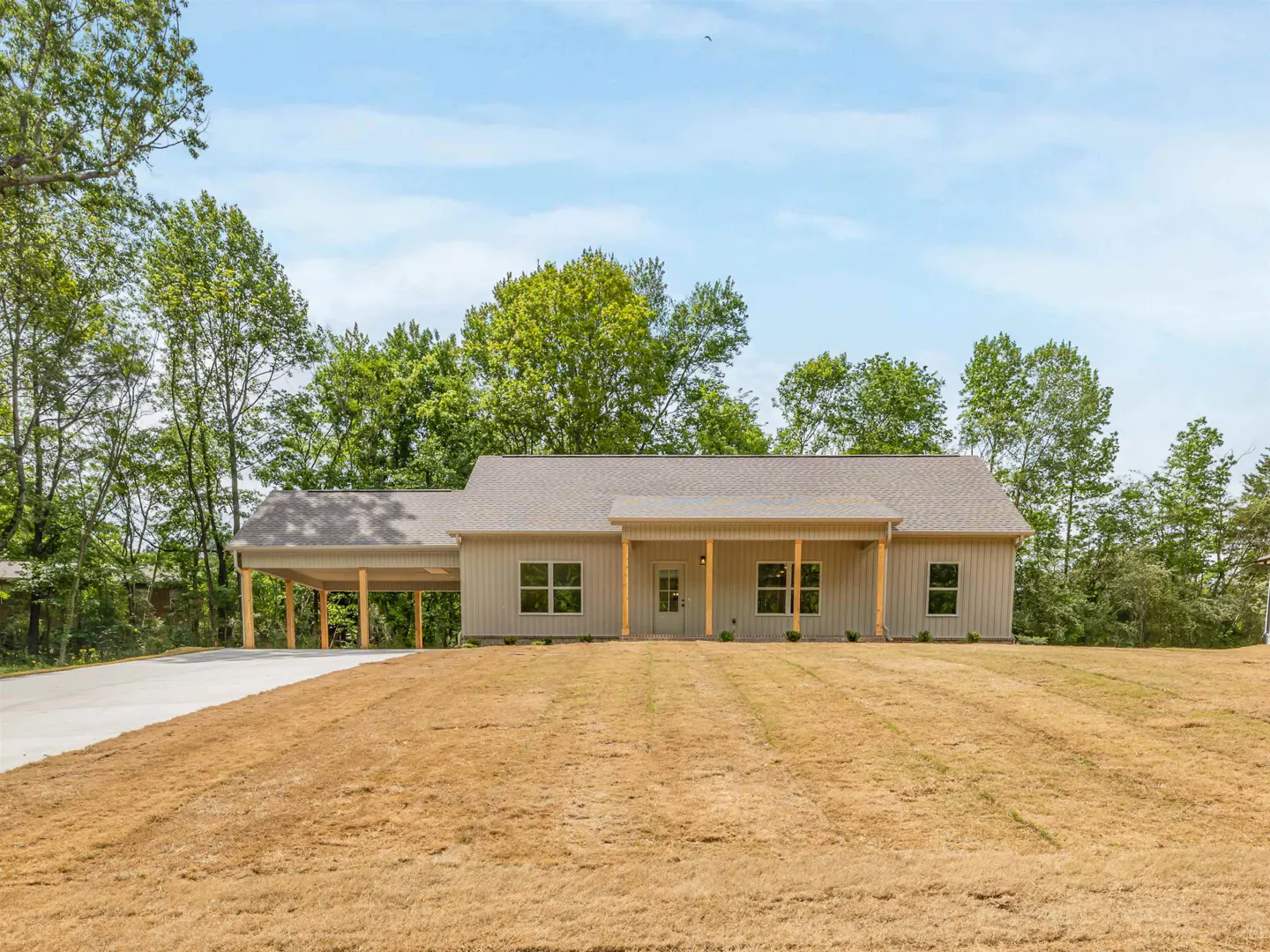 Tan house with a carport and a porch, surrounded by trees and a large lawn under a blue sky.