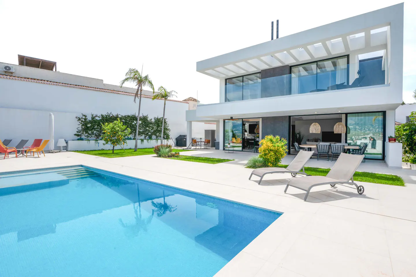 Modern two-story white house with a blue swimming pool, two lounge chairs, palm trees, and a white wall in the background.
