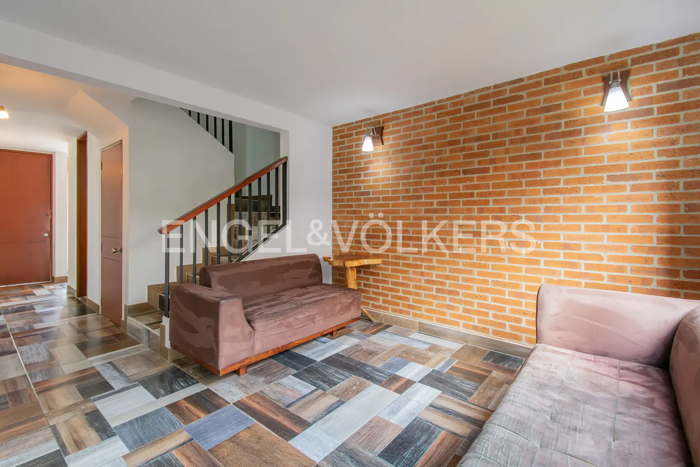 Living room with brick wall, brown sofa, and patterned tile floor. Stairs and Engel & Völkers logo in the background.