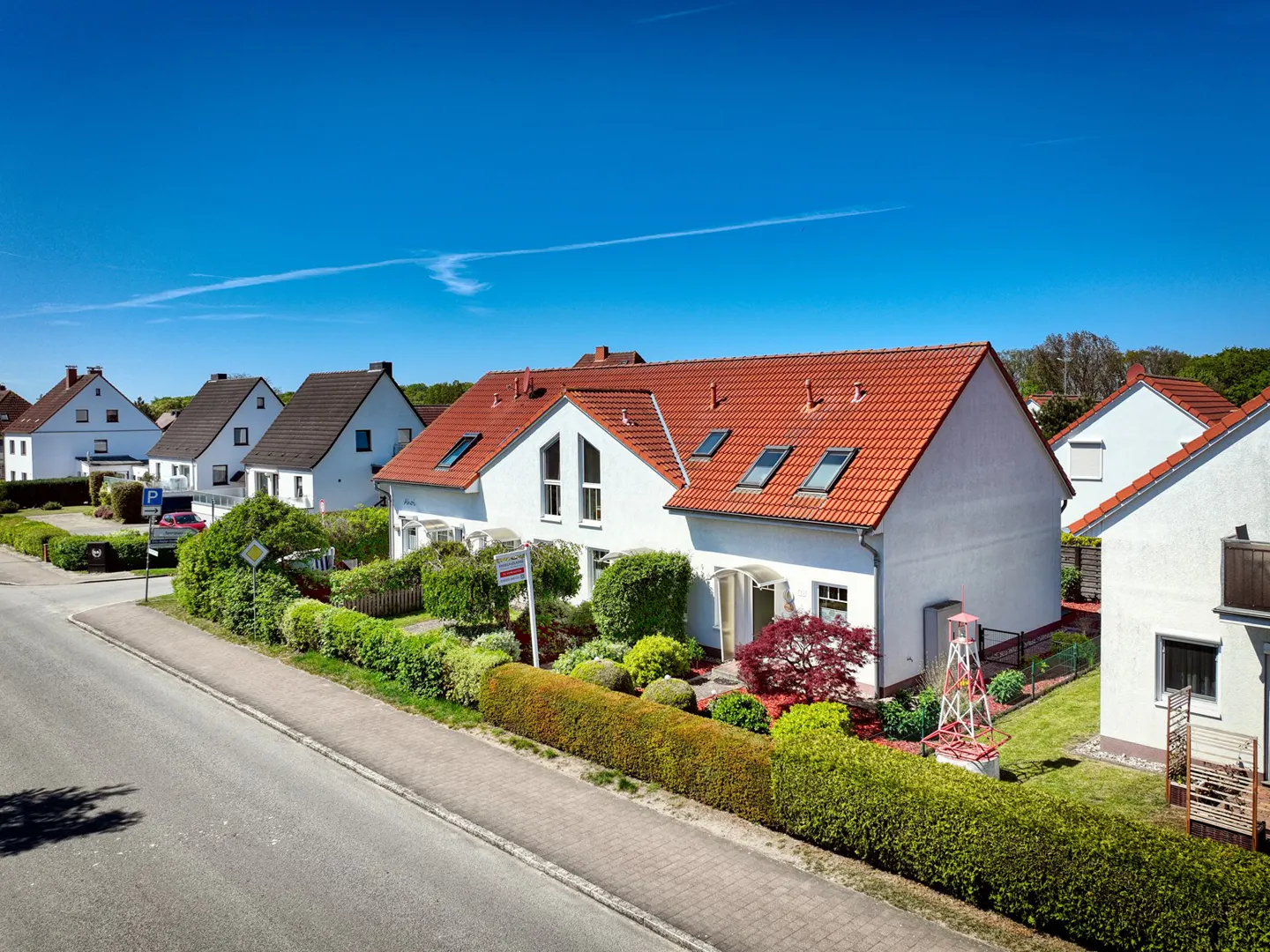 A white house with a red tile roof sits on a street with a blue sky above. A "For Sale" sign is in the front yard.