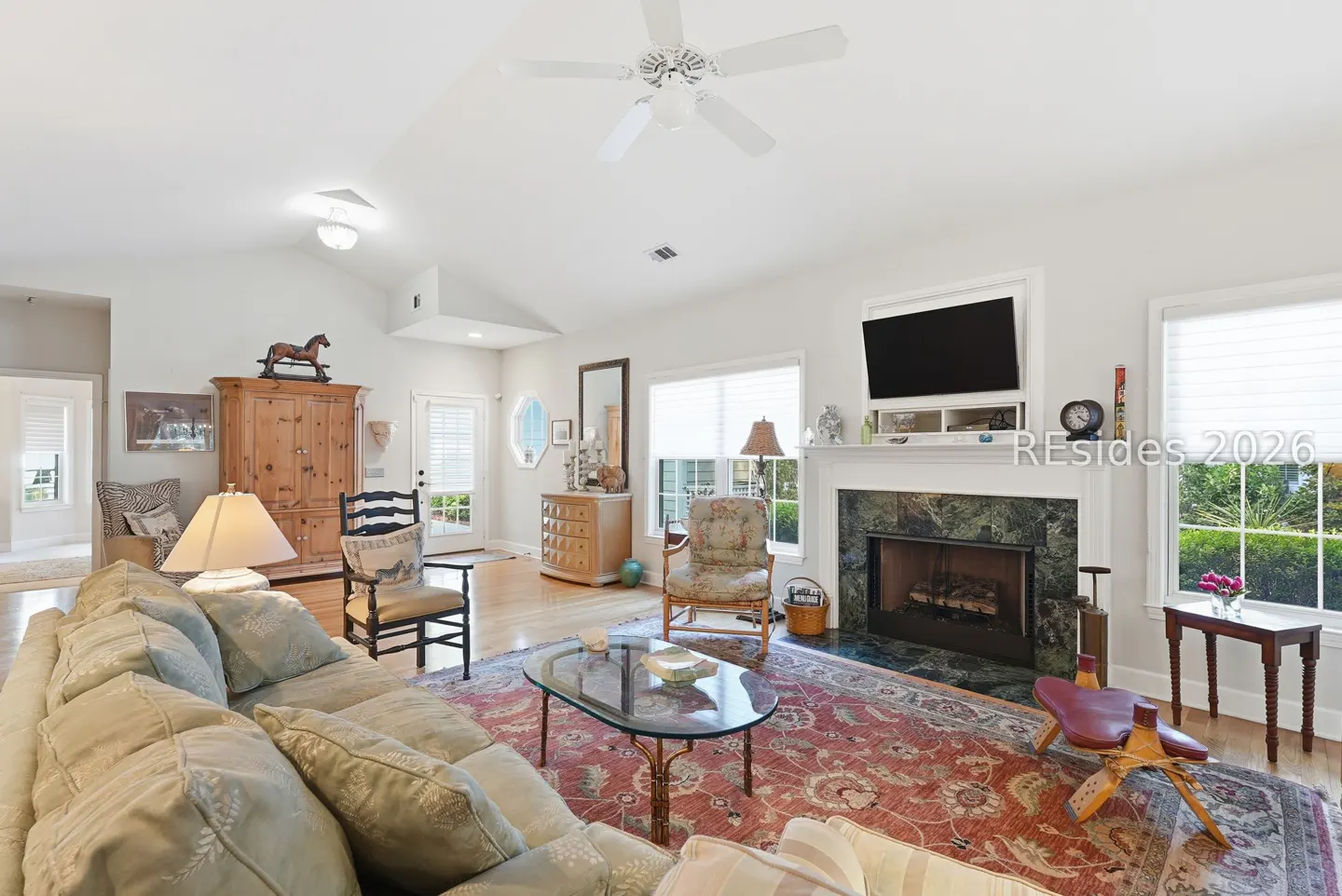 Bright living room with a large sofa, fireplace, TV, and a red patterned rug. A ceiling fan hangs above.