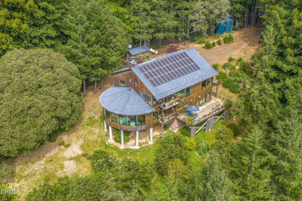 Aerial view of a wood-sided house with solar panels on the roof, surrounded by lush green trees. A round, glass-walled room is supported by white pillars.
