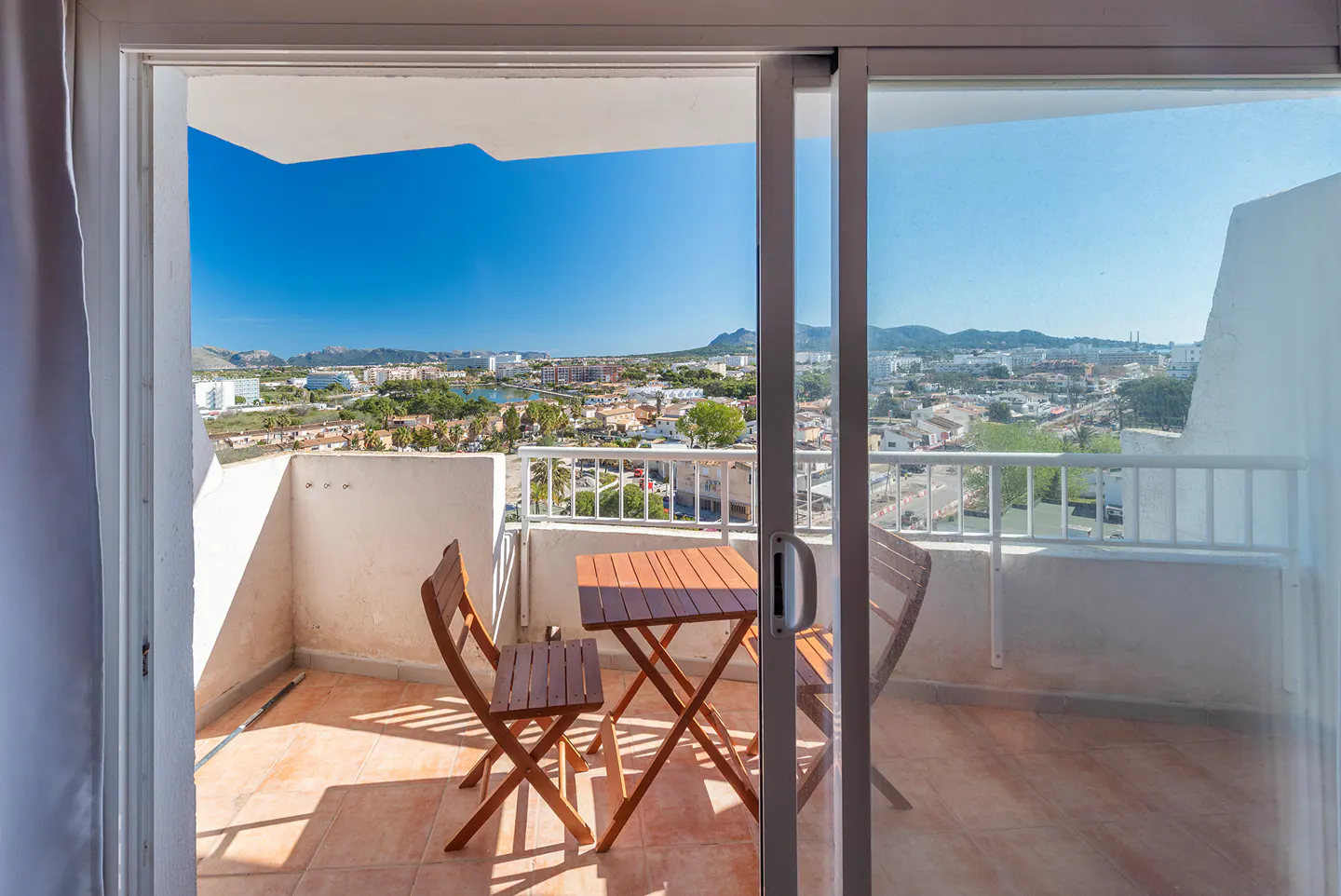Balcony with a brown table and two chairs, viewed through sliding glass doors. Cityscape and blue sky in the background.