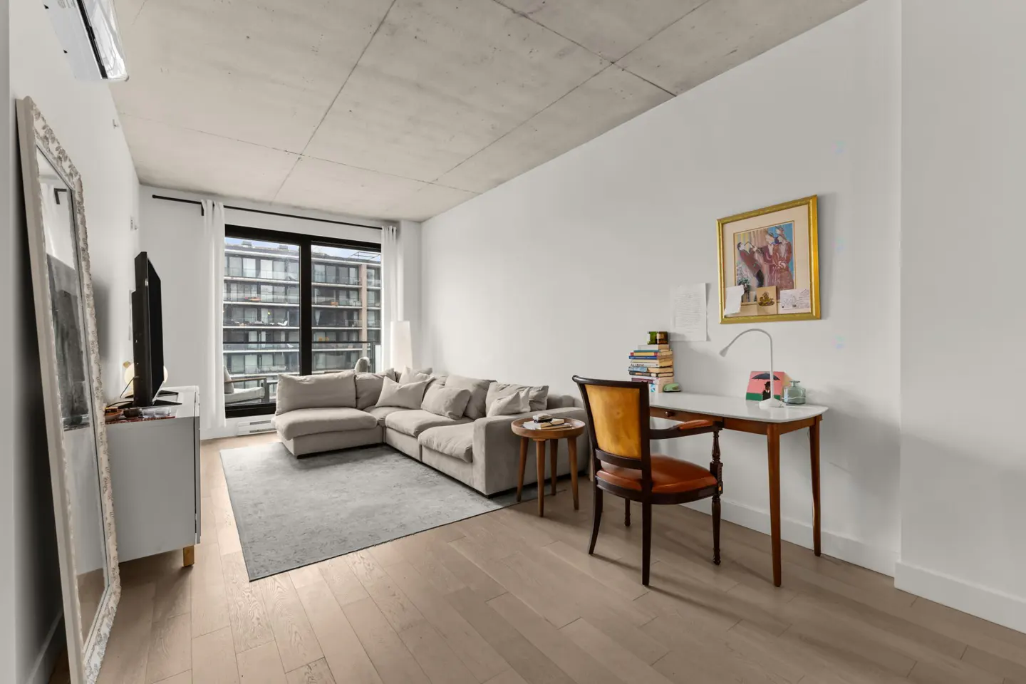 Living room with a gray sectional sofa, desk, and large window. Hardwood floors and white walls create a bright, modern space.