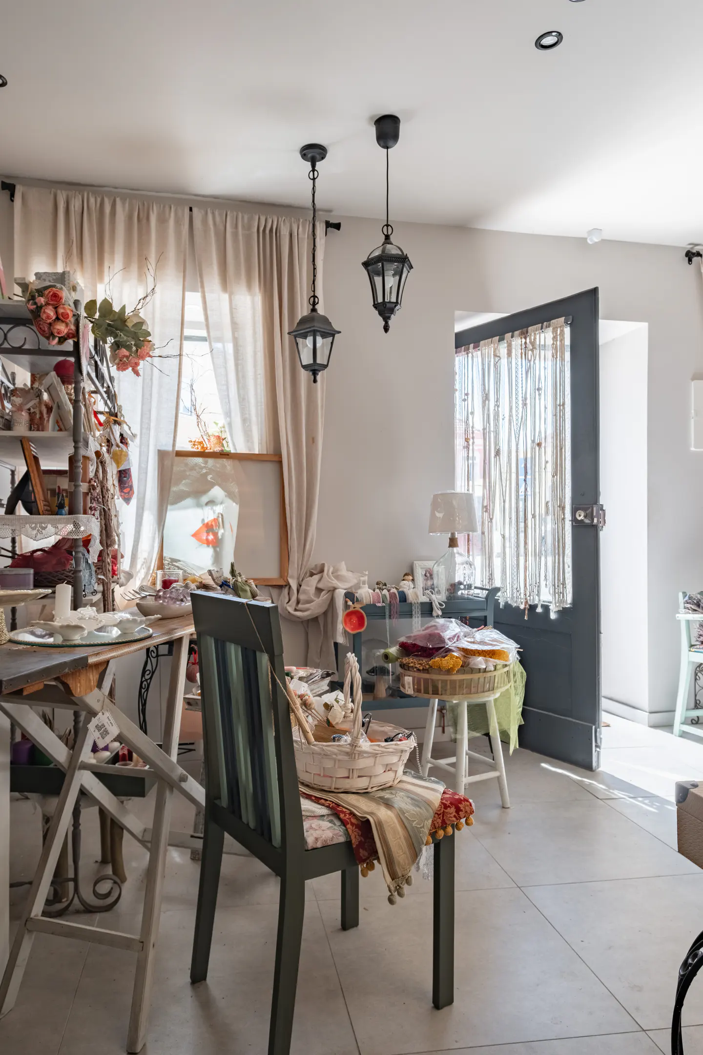 Bright, eclectic room with tables, chairs, and shelves filled with art supplies and decorative items. Two black pendant lights hang from the ceiling.