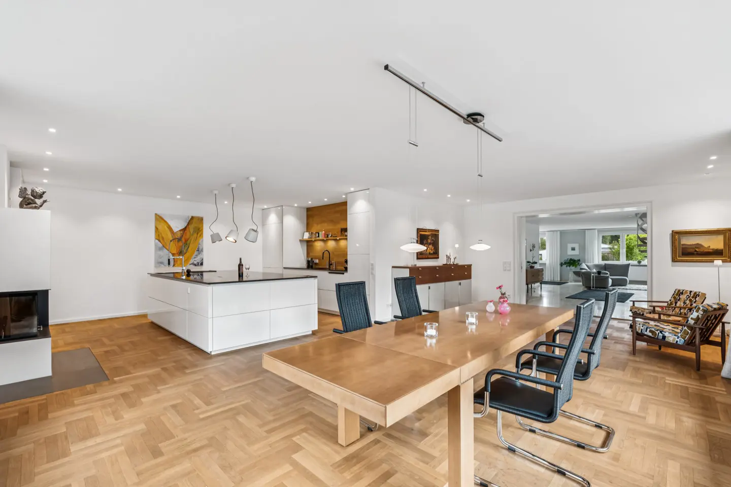 Open-concept living space with a kitchen island, dining table with six chairs, and a fireplace. Hardwood floors and white walls.