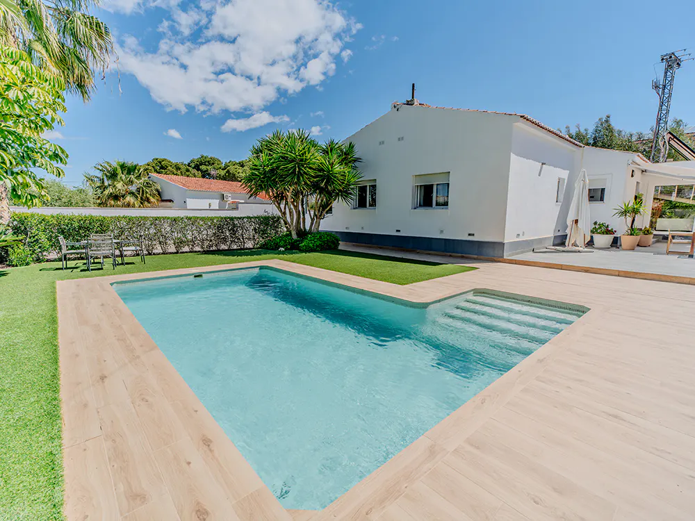A backyard pool with light blue water and wooden deck, next to a white house with green lawn and trees.