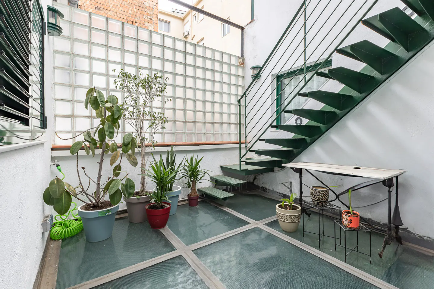 Outdoor patio with glass block wall, green metal stairs, and potted plants. A small table sits under the stairs.