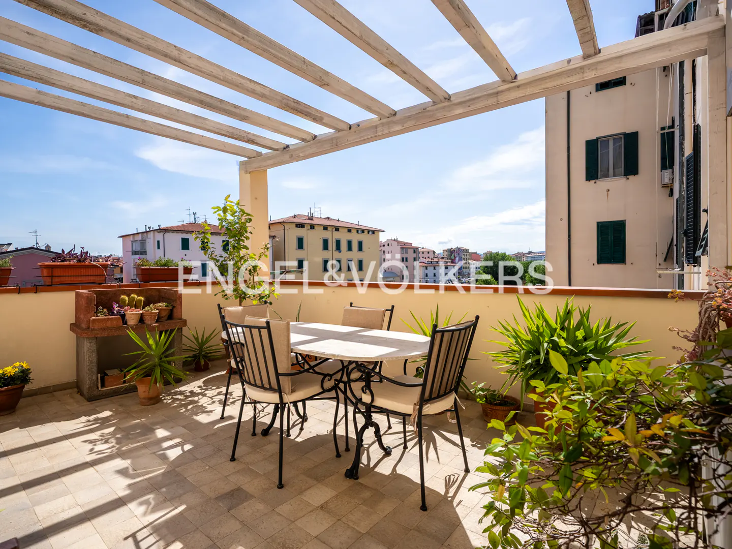 Outdoor terrace with a white table and four chairs, plants, and a pergola. City buildings are visible in the background.
