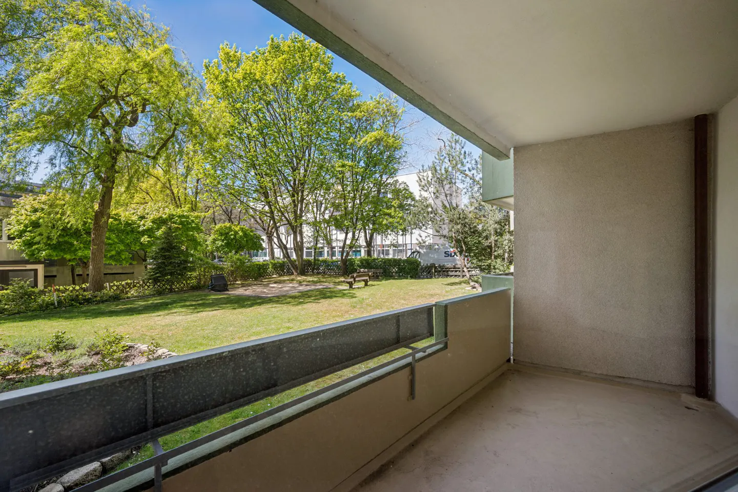 View from a balcony overlooking a green lawn with trees and a bench. The balcony has a metal railing and a concrete floor.
