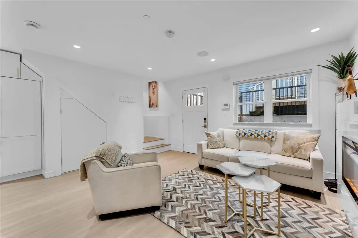 Bright living room with white walls, light wood floors, and a herringbone rug. A cream sofa and chair sit near marble-topped tables. Stairs lead up to a white door.