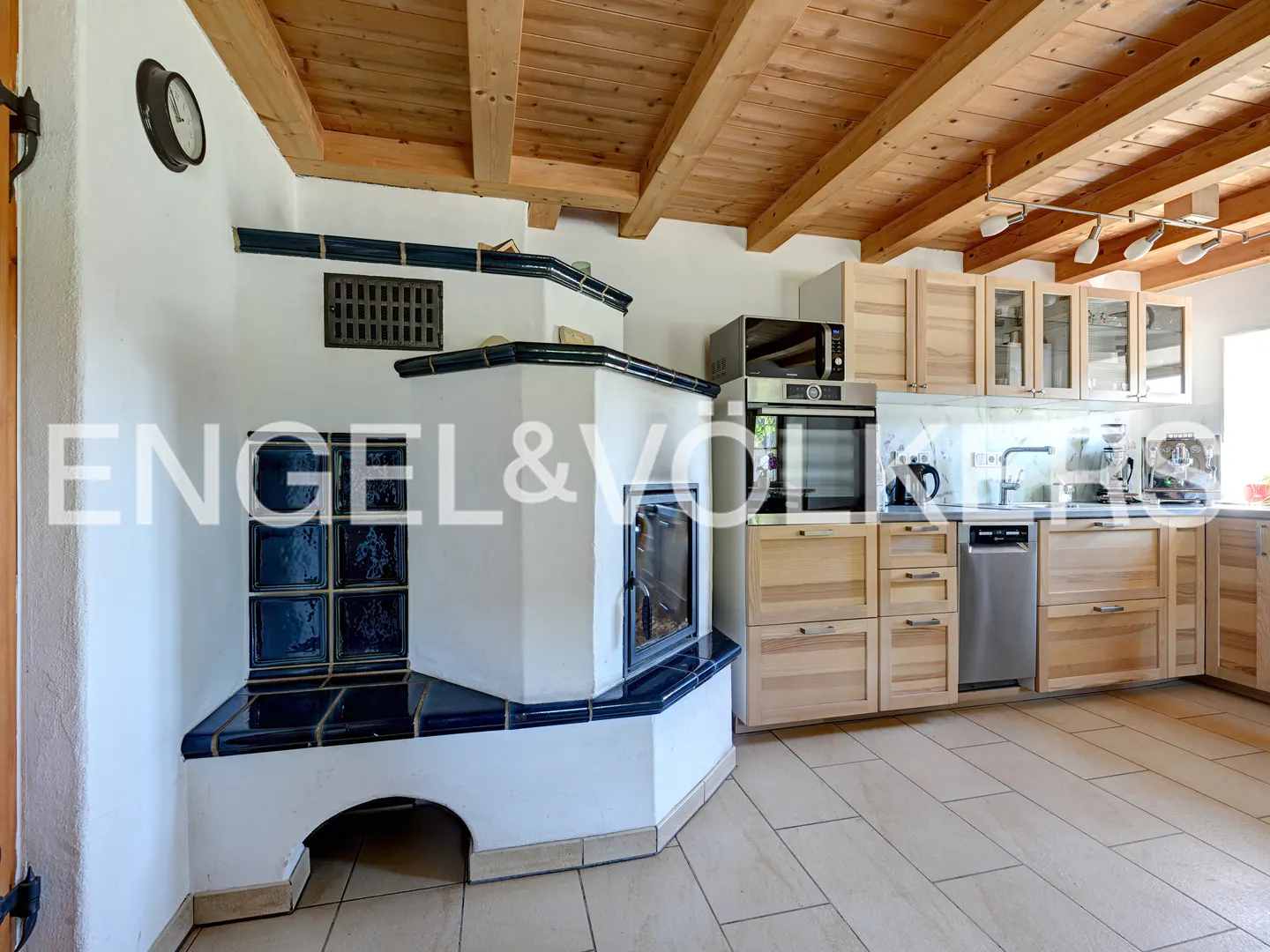 A kitchen with a white tiled stove, blue trim, and light wood cabinets under a wood-beamed ceiling.