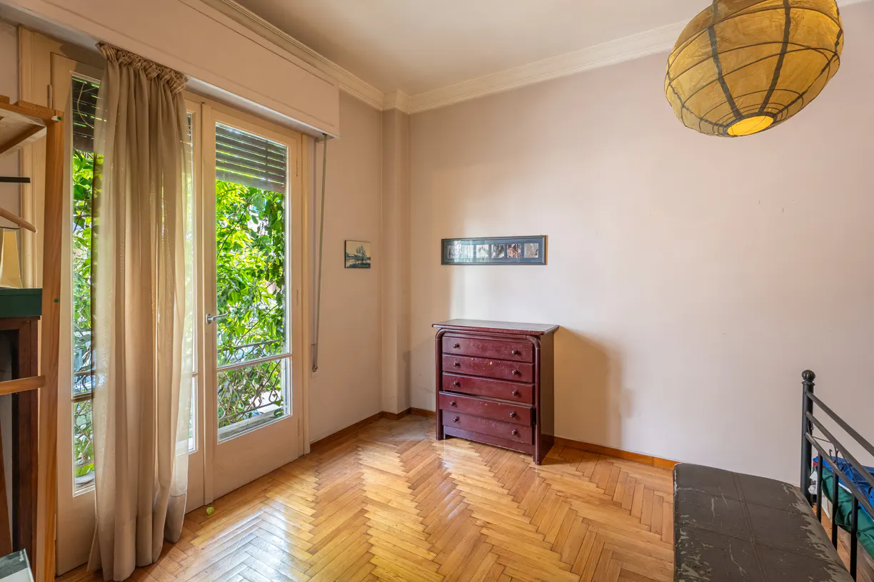 Bedroom with herringbone wood floors, a red dresser, and a window with sheer curtains overlooking green foliage. A paper lantern hangs from the ceiling.
