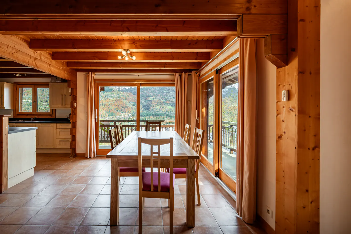 Interior of a wood cabin with a dining table, chairs with red cushions, and sliding glass doors to a balcony with a mountain view.