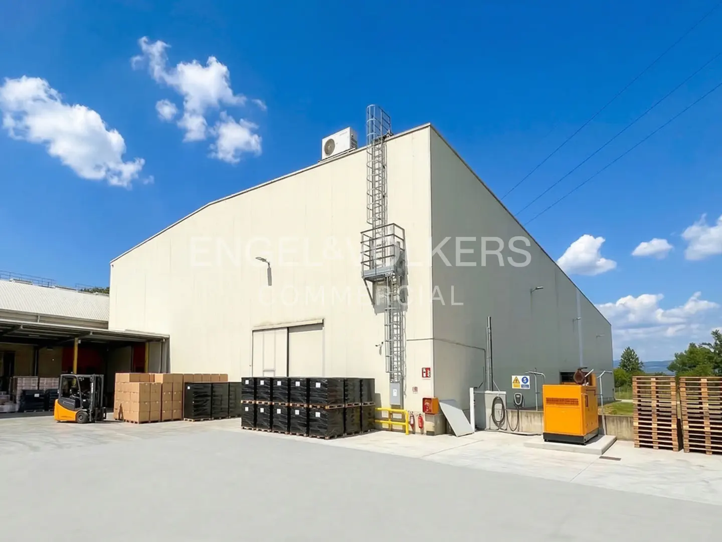 Exterior view of a large, light beige warehouse with a forklift, pallets, and a blue sky.