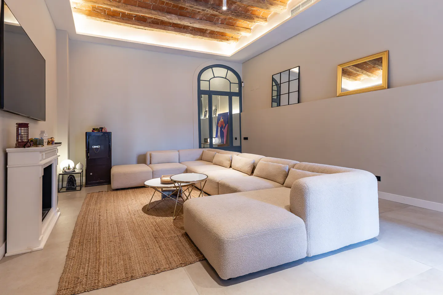 Living room with a large beige sectional sofa, round coffee tables, and a jute rug. A white fireplace and TV are on the left. Brick ceiling and arched doorway in the background.