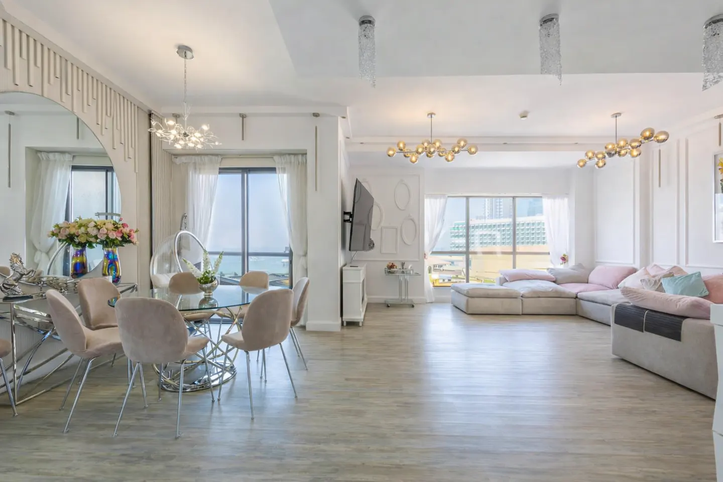 Bright, open-concept living space with dining table, plush sofa, and large windows overlooking the ocean. White walls and light wood floors.