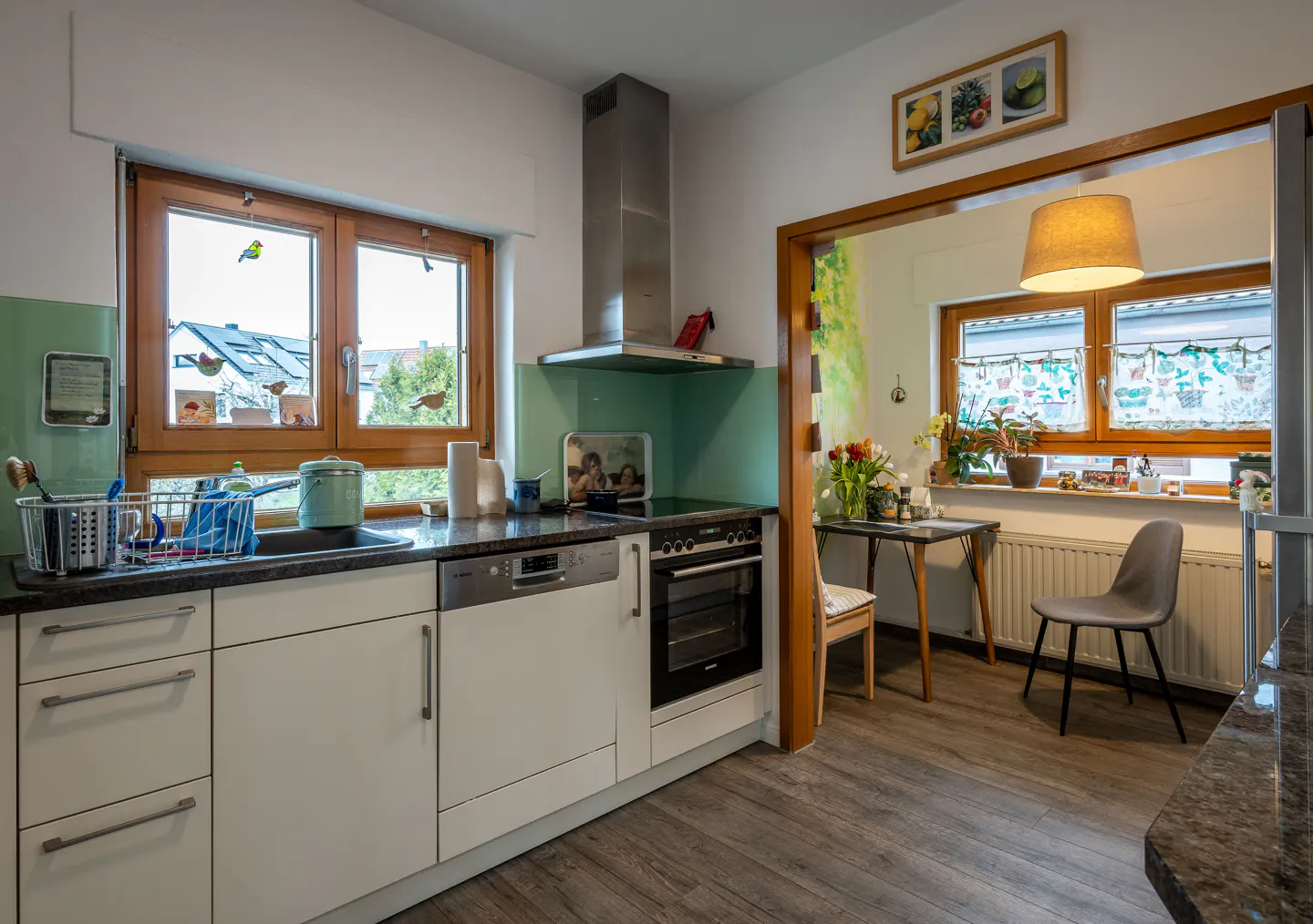 A bright kitchen with white cabinets, a stainless steel range hood, and a window over the sink. An open doorway leads to a dining area with a table and chair.