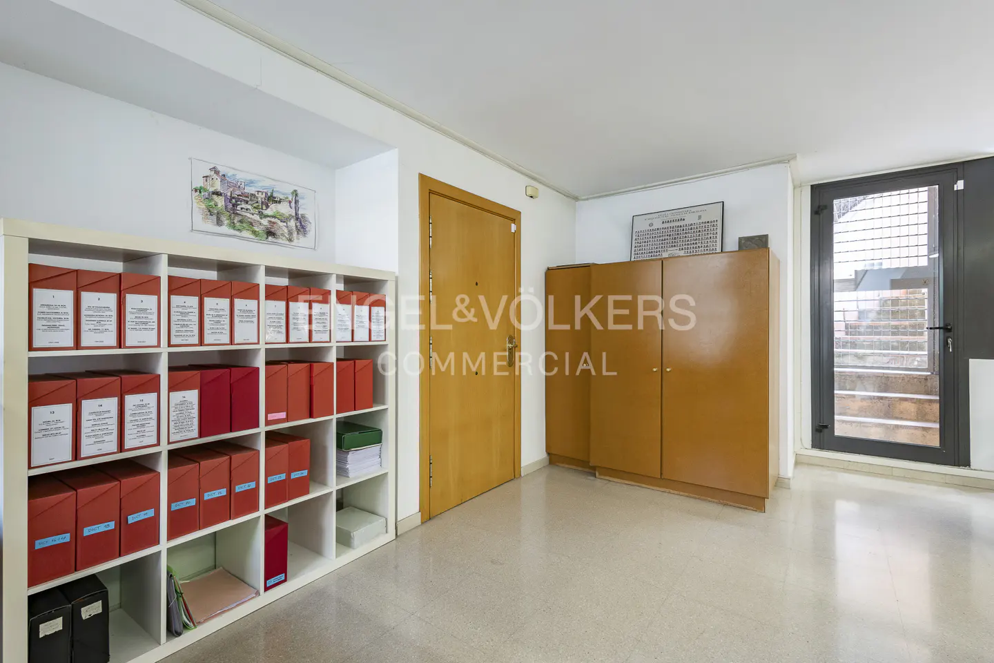 Office interior with a white shelving unit filled with red file boxes, a wooden door, and a brown cabinet. A black door with glass panes is on the right.