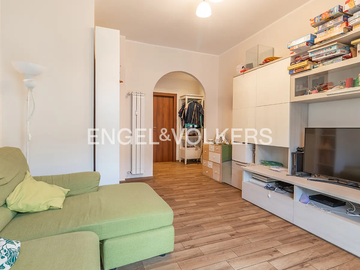 Living room with a green sofa, light wood floors, and white built-in cabinets with a TV. A hallway leads to a wooden door.