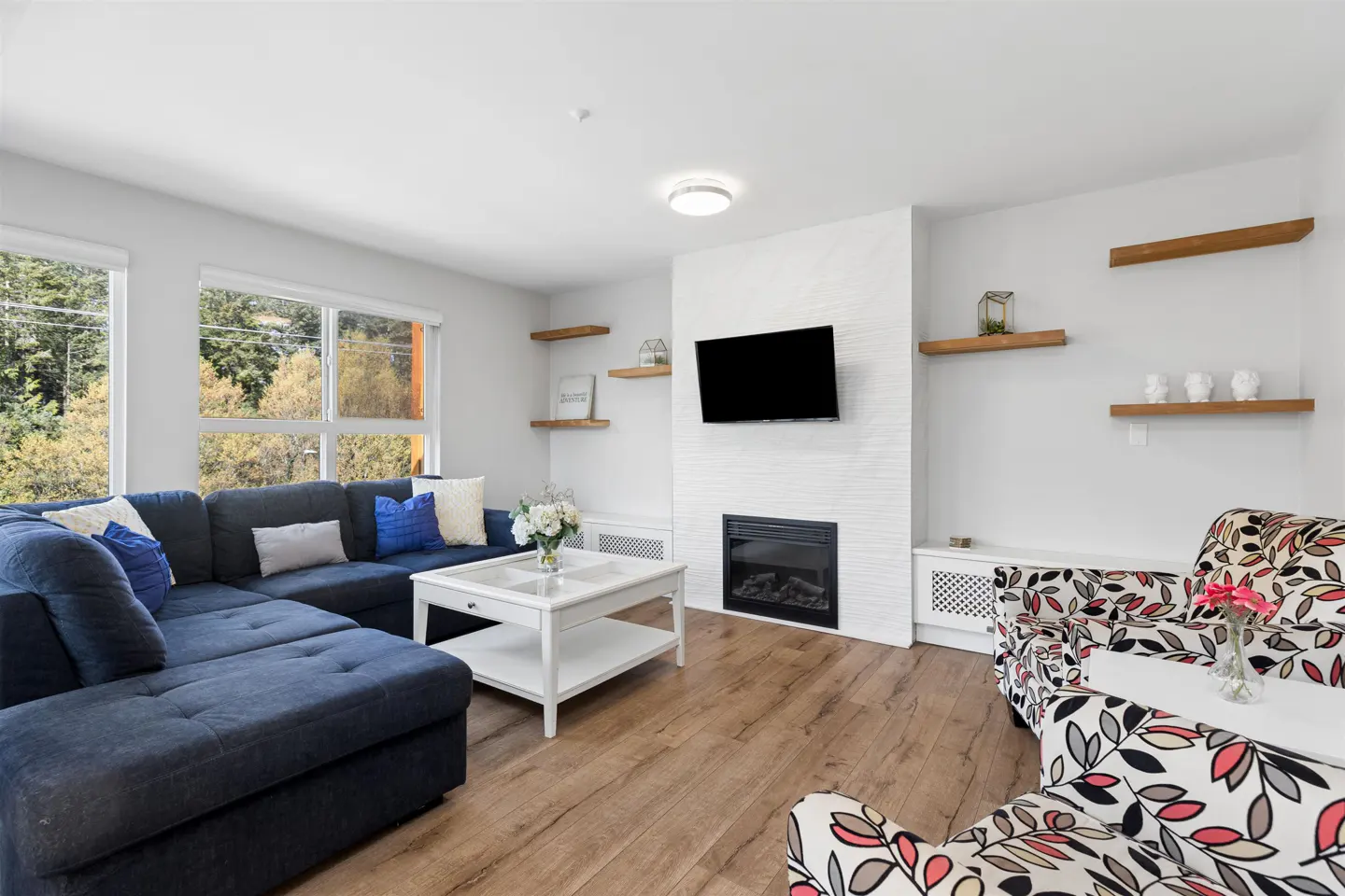 Bright living room with a blue sectional sofa, white coffee table, fireplace, TV, and floral armchairs on a wood floor.